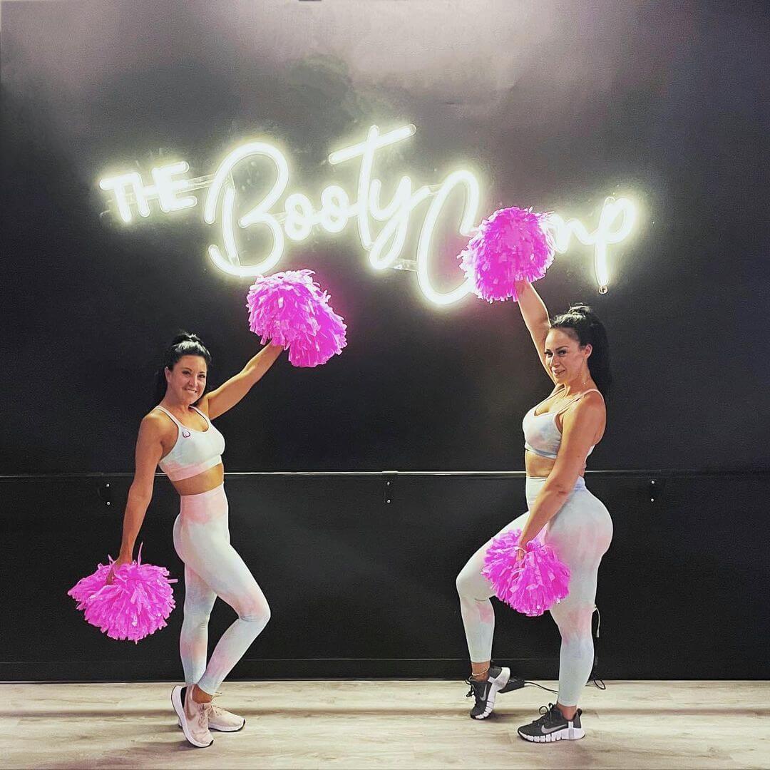 2 cheerleaders with The Booty Camp white neon sign behind them