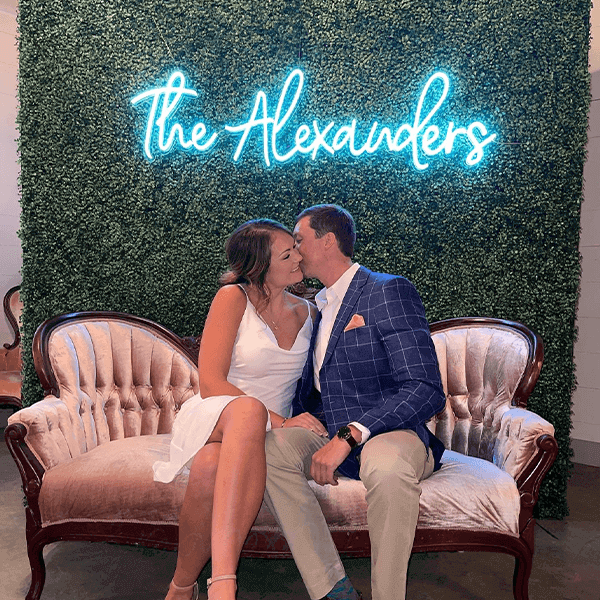 Couple sitting in front of the Alexanders' last name turquoise wedding event neon sign 